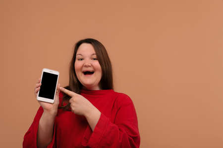 A smiling girl points her hand at the blank screen of her phone. Beige background.の写真素材