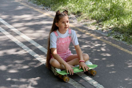 A cute beautiful girl is sitting on the track with a skateboard in her hands. Summer holidays. Hot summer day.の写真素材
