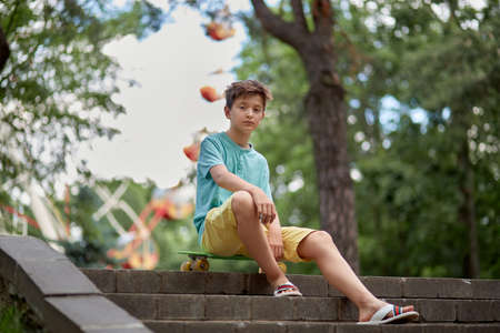 A child boy is sitting in an amusement park on the steps with a skateboard in his hands. Summer walks. Blurred background.の写真素材