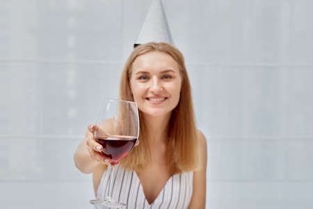 Birthday celebration, holding a glass with red wine in his hand. Light background. Woman in a festive cap.の写真素材