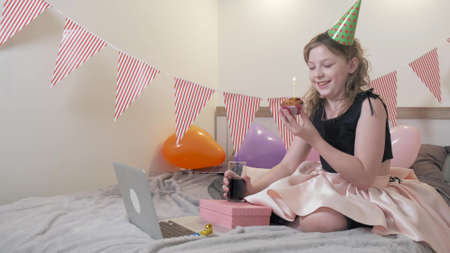 The girl will blow out the candles on the birthday cake. Beautiful decorated room with flags and balloons. Communication using social networks and video communication.の写真素材