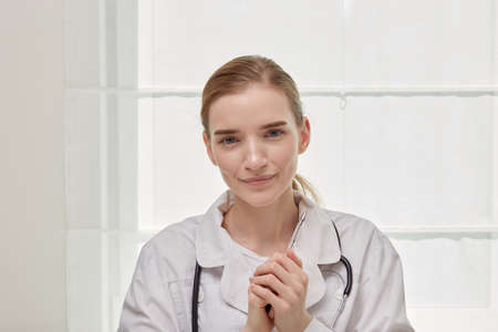 Portrait of a young cute female doctor in a white medical coat. White background. Hands clasped together.の写真素材