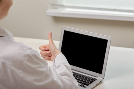 A female doctor shows a thumbs-up gesture and looks at a black laptop monitor. Online consultation.