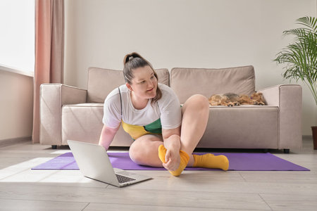 An overweight woman does sports at home online. A pet, a small dog, is sleeping on the sofa.の写真素材