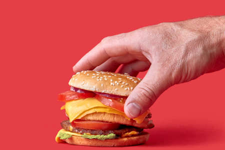 Hand half of the buns with sesame seeds on a big juicy Burger. Burger with cheese, vegetables and two cutlets. Red background.の写真素材