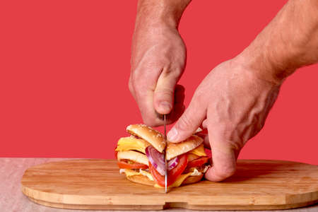 A mans hand cuts a large Burger on a light wooden Board with a knife. Red background.の写真素材