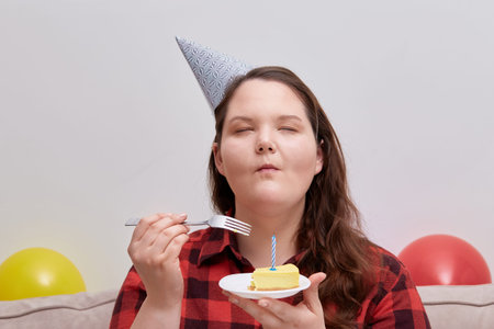A young woman enjoys a delicious birthday cake with her eyes closed. Plus size model. Colorful...