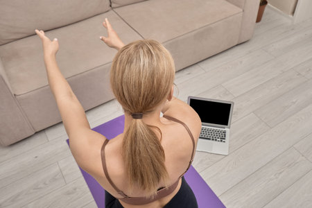 Back view of a young woman looking at a black laptop screen, learning yoga poses online. Sports for...