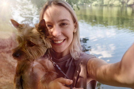 Smiling happy young blonde woman takes a selfie with her beloved dog in the background of the lake. Bright Sunny morning.の写真素材