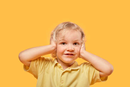Portrait of a beautiful boy with curly blond hair on a yellow background. Your ears are covered with your hands. Looks at the camera.の写真素材