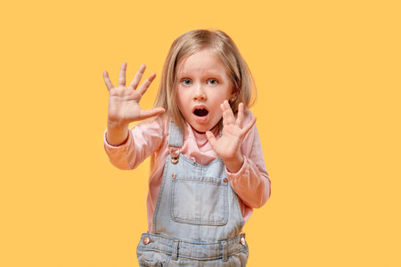 The frightened little girl puts her hands out and defends herself. Yellow isolated background.