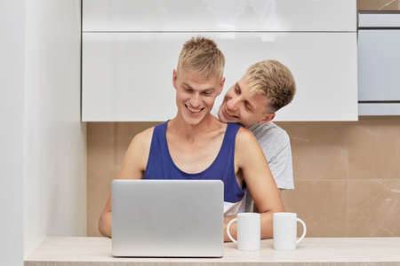 Cute beautiful loving couple of two LGBT men in the morning in the kitchen. There is an open laptop on the table.の写真素材