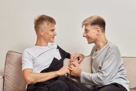 Happy LGBT couple of two men communicate at home sitting on the couch. Light wall on the background.の写真素材
