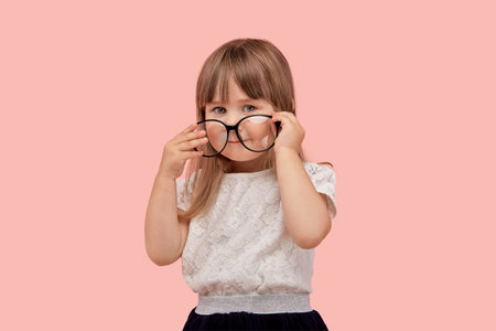 The girl puts on large glasses for vision. Poor eyesight in a child. He touches the glasses with his hands. Pink isolated background.の写真素材