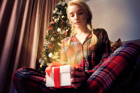 Beautiful young woman in plaid pajamas opens a gift. From the box shines a Golden, yellow light and fly small sparks, sparkles. Christmas and new year celebrations.の写真素材