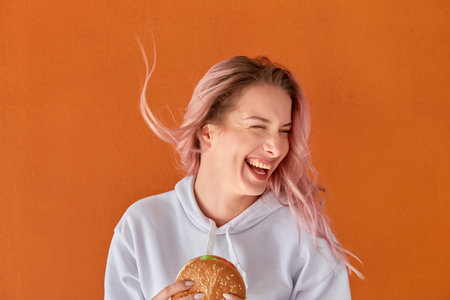 A happy woman holds a large hamburger in her hand and laughs. A woman on a diet. The concept of harmful nutrition. Orange background.の写真素材
