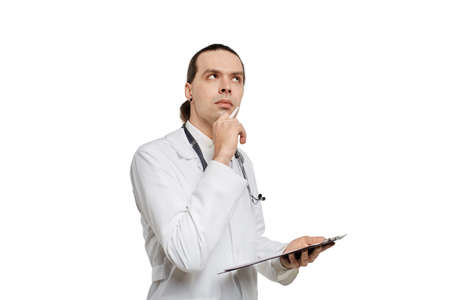 A young, handsome male doctor with a clipboard in his hands thinks and looks away. White isolated background.の写真素材