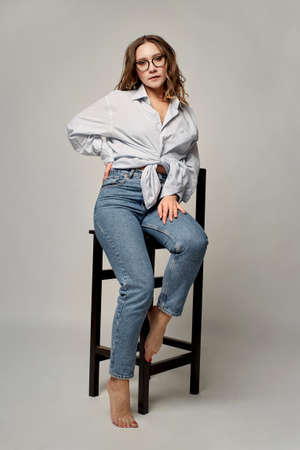 A young, beautiful, stylish woman wearing black-rimmed glasses, a shirt, and jeans sits on a bar stool and looks at the camera. Photo in the studio.の写真素材