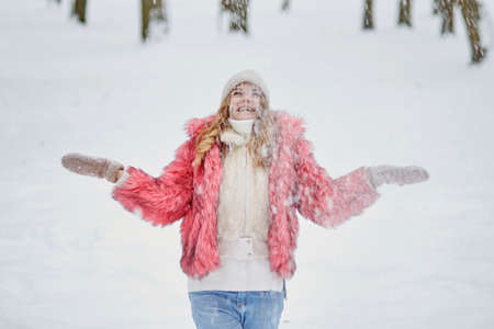 A beautiful young woman in a winter park is standing under a snowfall, a lot of snow. Pink fur coat and white knitted hat. A cold, snowy winter.の写真素材