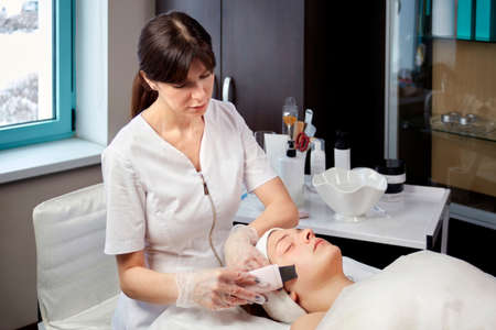 A cosmetologist in a medical gown does a facial cleansing procedure for a client. Beauty salon. Professional skin care products.の写真素材