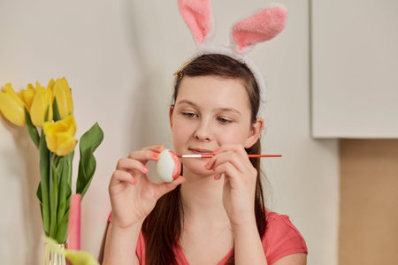 A brunette girl paints an Easter egg with red paint. There are yellow tulips on the table. The...