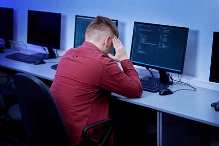A young man stands on his head while sitting at a computer in the office. Headache. The codes are displayed on the monitor.の写真素材