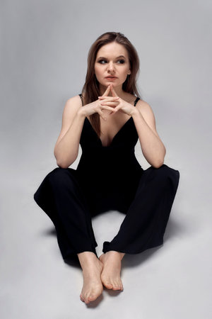 A young woman in a black jumpsuit sits on the floor barefoot and looks away. Black formal clothing. Business woman in black clothes.の写真素材