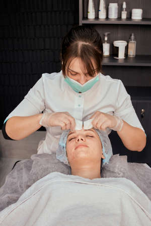A female cosmetologist removes acne with her hands on the face of a young beautiful woman. Professional beauty salon. Vertical photo.の写真素材