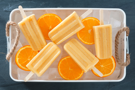 Homemade orange yogurt popsicles in a rustic ice filled tray with fresh fruit slicesの写真素材