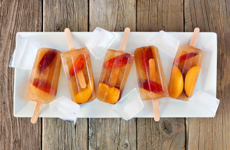 Peach iced tea popsicles with ice cubes on white plate and rustic wood backgroundの写真素材