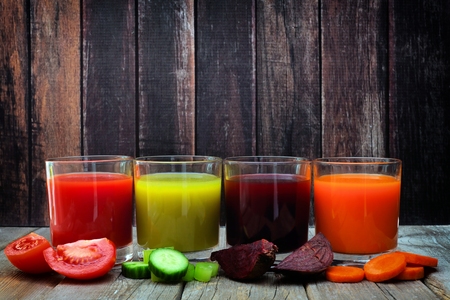 Four glasses of healthy vegetable juice with scattered vegetables and a dark wood backgroundの写真素材