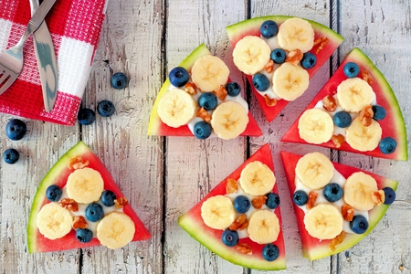 Sliced, juicy watermelon pizza with bananas, blueberries, nuts and yogurt, overhead view on rustic white woodの写真素材
