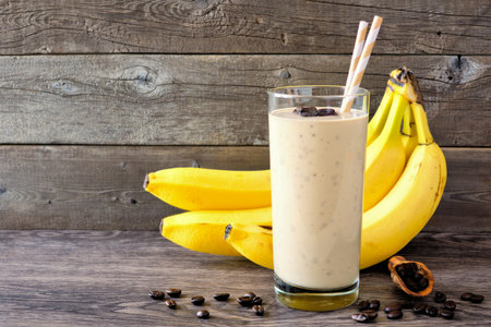 Coffee, banana smoothie in a tall glass with coffee beans and bananas in background. Side view, against a rustic wood background.の写真素材