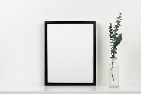 Mock up black frame with vase of branches on a shelf or desk. White shelf and wall. Portrait frame orientation.の写真素材