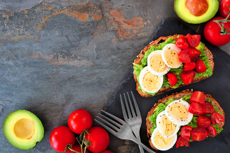 Healthy avocado toasts with eggs and tomatoes. Flat lay, corner border over a dark stone background.の写真素材
