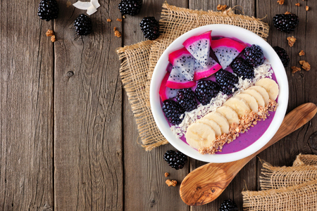Blackberry smoothie bowl with coconut, bananas, dragon fruit and granola. Above view corner border on a wood background.の写真素材