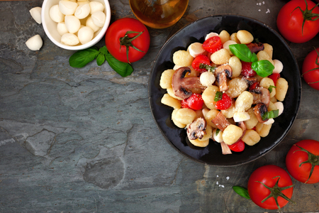 Gnocchi with tomatoes, mozzarella, mushrooms and basil, top view corner border over a dark stone background with copy space.の写真素材
