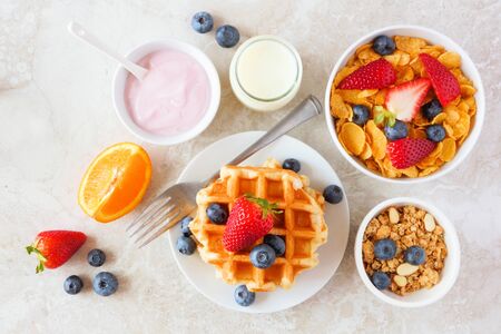 Breakfast food table scene of fruits, cereal, waffles, yogurt and milk, top view over a bright stone backgroundの写真素材