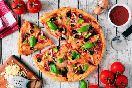 Homemade pizza with pepperoni, vegetables and basil, top view table scene with cut slices over white woodの写真素材