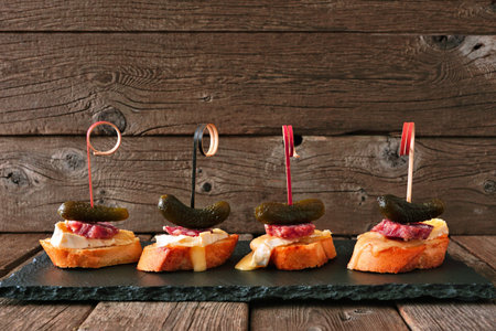 Crostini appetizer skewers with baked brie, sausage and pickles, serving platter against rustic wood