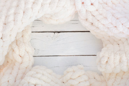 Cozy cream colored knitted thick wool blanket, top view frame on a white wood background