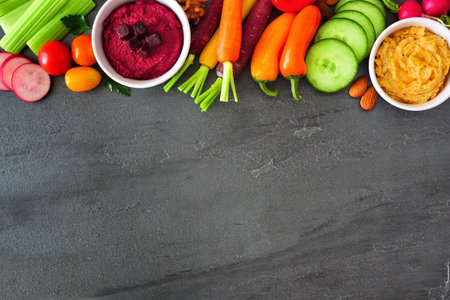 Variety of fresh vegetables and hummus, above view top border on a dark slate background