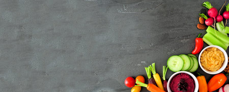 Assortment of fresh vegetables and hummus, above view corner border on a slate banner background