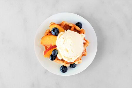 Dessert waffle with vanilla ice cream, peaches and blueberries, top view on white marbleの写真素材