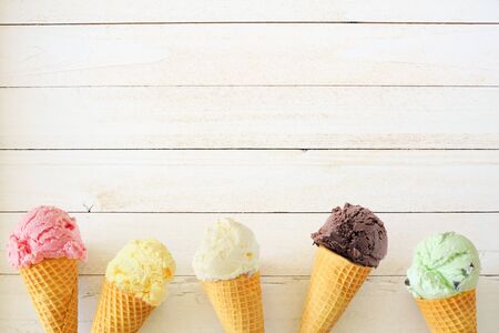 Ice cream cone bottom border with a variety of flavors, top down view over a white wood backgroundの写真素材