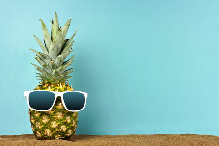 Pineapple with sunglasses in sand against a teal background. Summer beach concept.