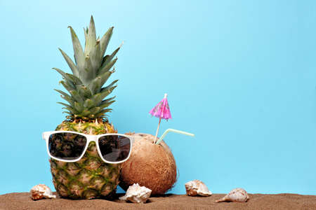 Pineapple with sunglasses in sand with coconut drink against a blue background. Summer beach concept.の写真素材