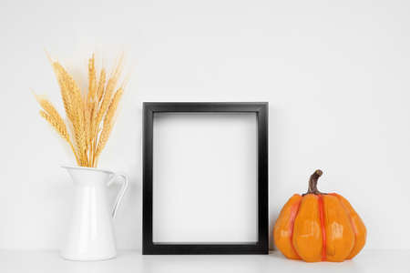 Mock up black frame with fall wheat and pumpkin decor on a white shelf. Autumn concept. Portrait frame against a white wall.の写真素材