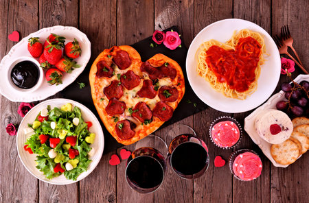 Home cooked Valentines Day dinner. Above view table setting on a dark wood background. Heart shaped pizza, pasta, wine, cheese plate and desserts.の写真素材