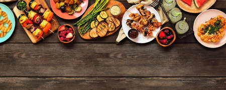 Healthy plant based summer bbq top border. Above view over a dark wood banner background. Grilled fruit and vegetables, skewers, cauliflower steak and vegetarian sides. Copy space.の写真素材